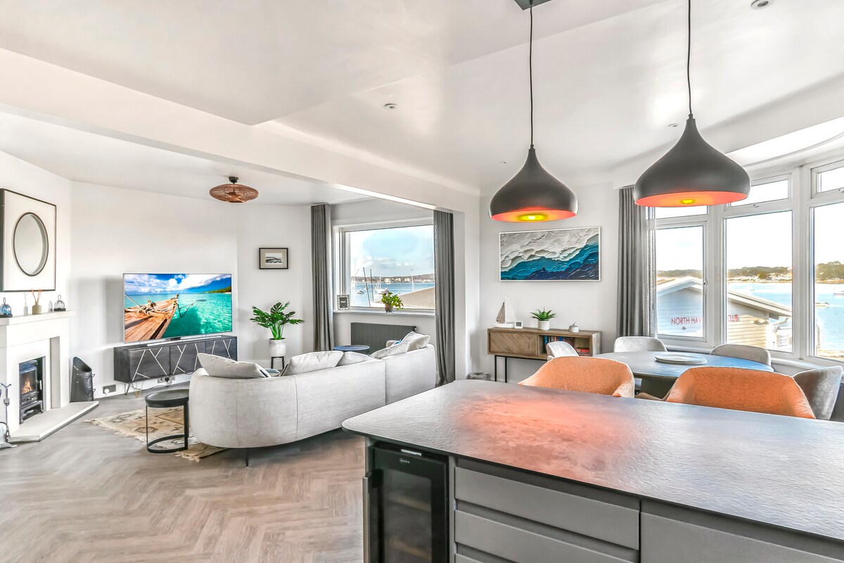 An open-plan living area is shown, featuring a comfortable grey sofa and a large TV mounted on the wall. The room is illuminated by modern pendant lights, with large windows offering views of the harbor. A dining table and stylish decor enhance the welcoming ambiance.