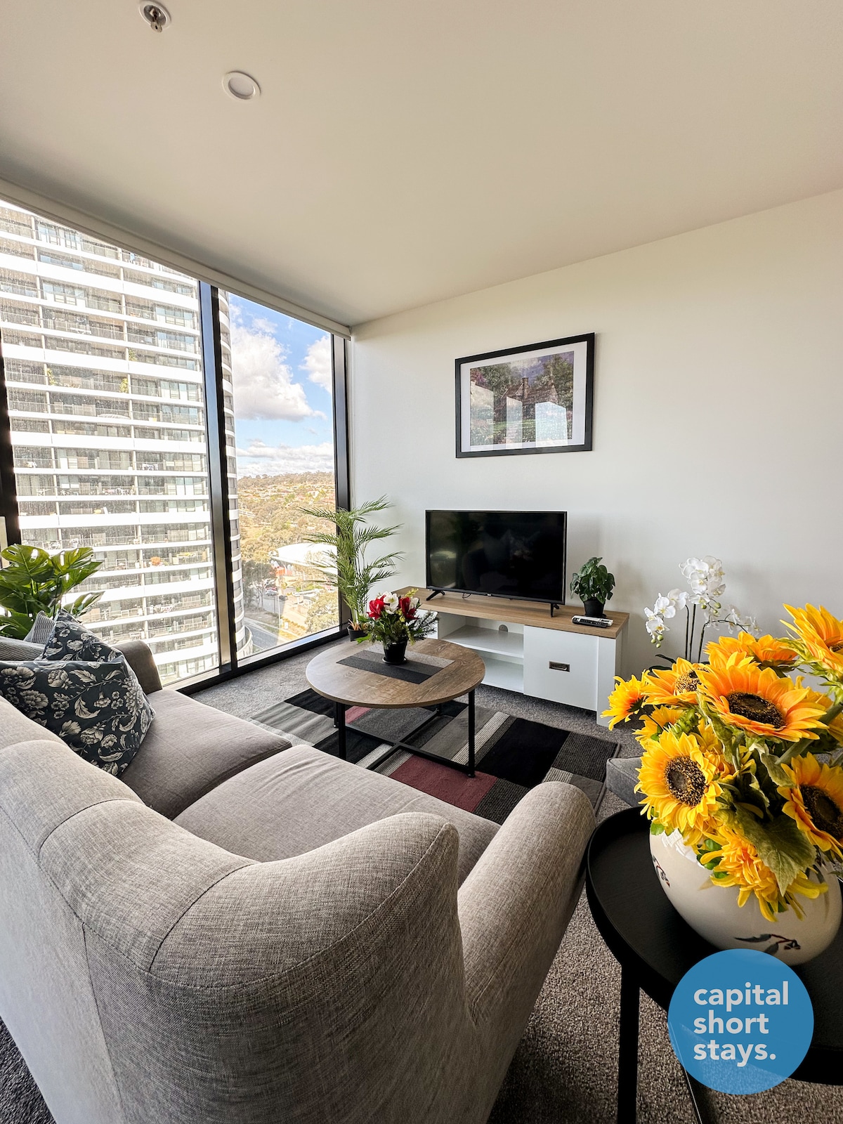 The bright living area features large floor-to-ceiling windows that showcase sweeping views of the surroundings. A plush gray sofa rests beside a circular coffee table adorned with vibrant flowers. A smart TV is placed on a console against the wall, enhancing the welcoming atmosphere.