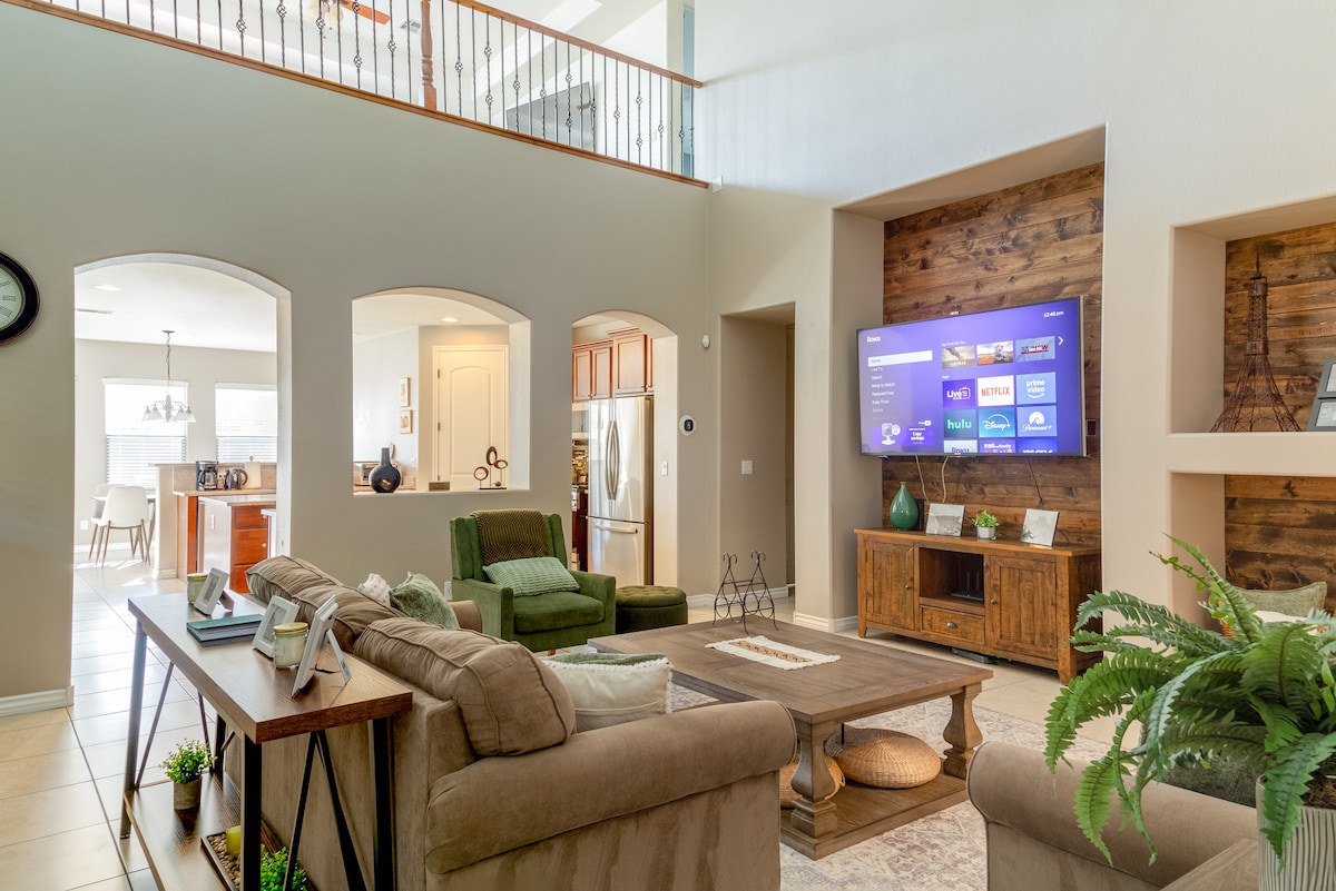 A spacious living area features a large, neutral-colored sectional sofa with an assortment of cushions. A wooden coffee table sits at the center, complemented by a decorative plant. A mounted TV screens entertainment options against a rustic wooden panel, while natural light streams through arched entryways.