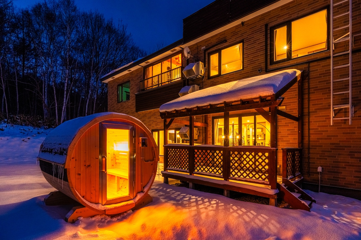 A stylish wooden sauna is positioned beside a deck area illuminated warmly in the evening. The exterior of the building is seen, with large windows showing a welcoming glow. Fresh snow covers the ground, enhancing the cozy atmosphere.