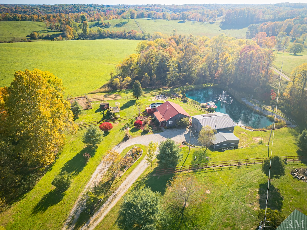 An aerial view showcases the charming cottage surrounded by lush greenery and colorful trees. A winding driveway leads to the home, while a serene pond is visible nearby, reflecting the natural beauty of the landscape.