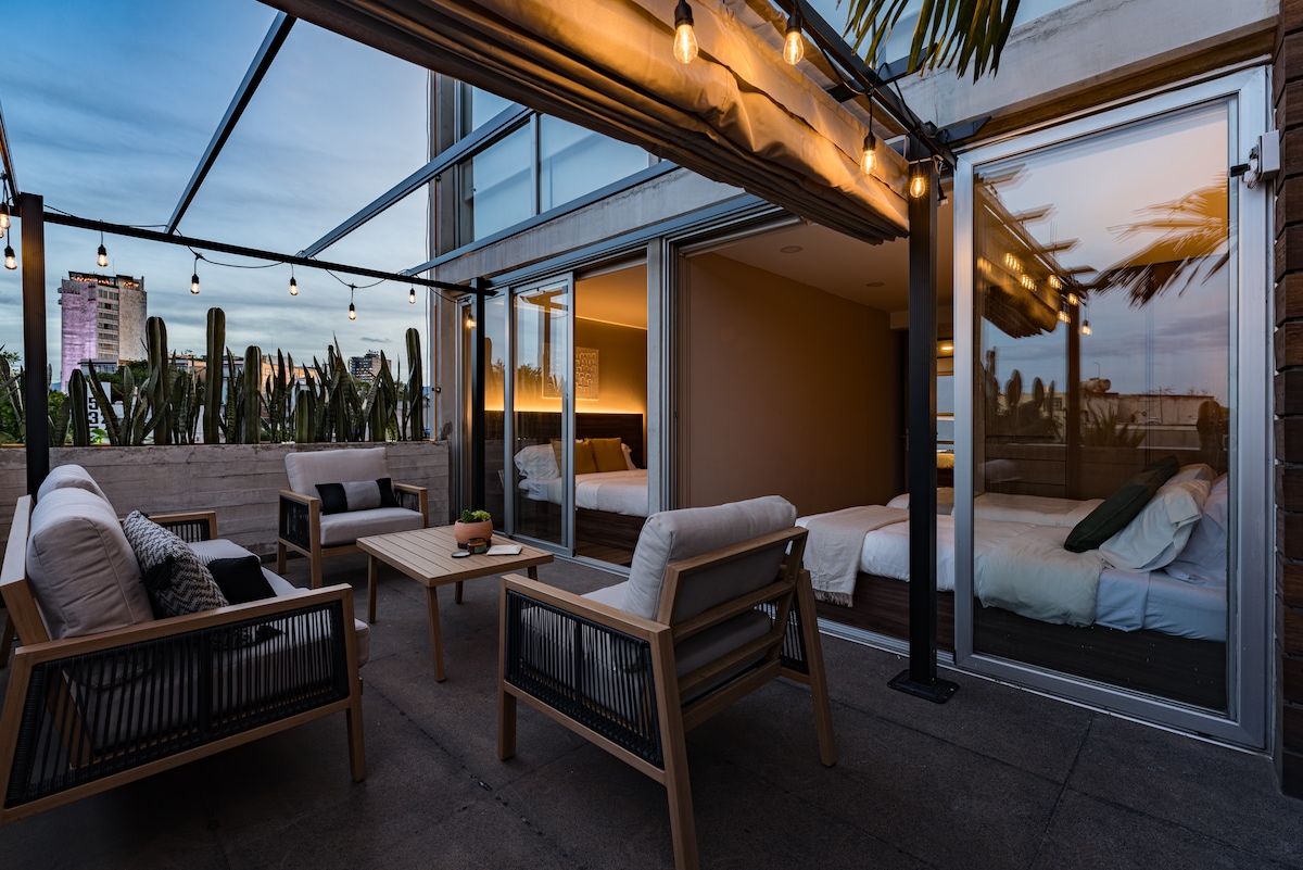 An inviting terrace is furnished with comfortable seating and a wooden table, surrounded by lush greenery. Soft lighting from overhead string lights creates a warm ambiance. Large glass doors provide a view of the interior bedrooms, blending outdoor and indoor spaces seamlessly.
