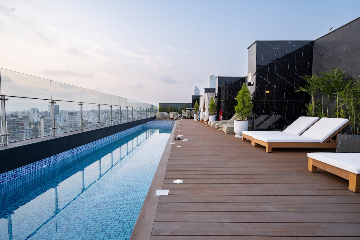 A rooftop pool area features a long, tiled pool extending alongside a wooden deck. Lounge chairs are positioned for relaxation, while planters add greenery to the space. The skyline view offers a subtle backdrop against a light blue sky.