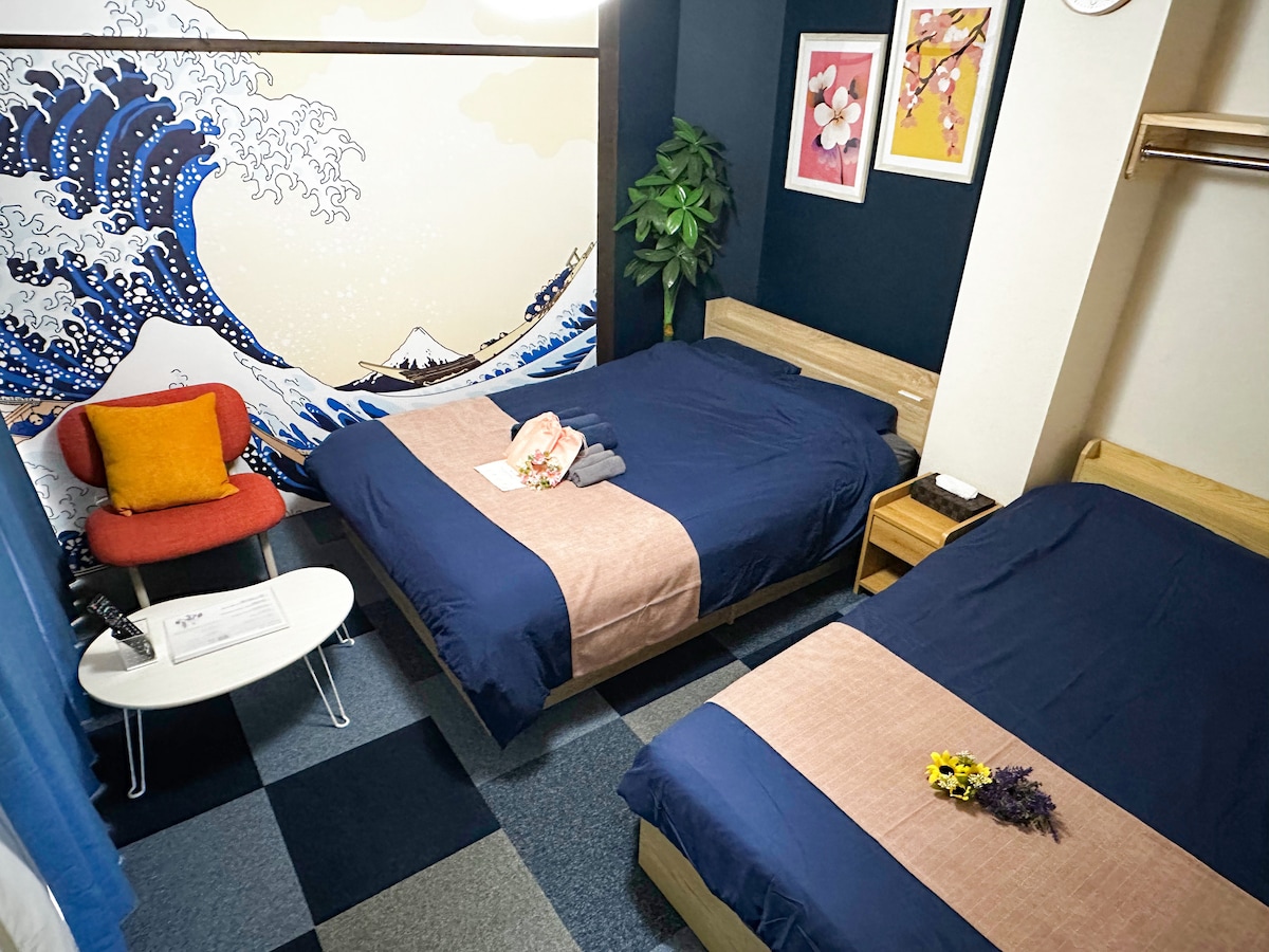 A room features two spacious beds with blue bedding draped over light wooden frames. A decorative wall displays a wave mural. A small table with a chair is positioned beside the beds. A plant adds a touch of greenery, complemented by framed artwork on the wall.