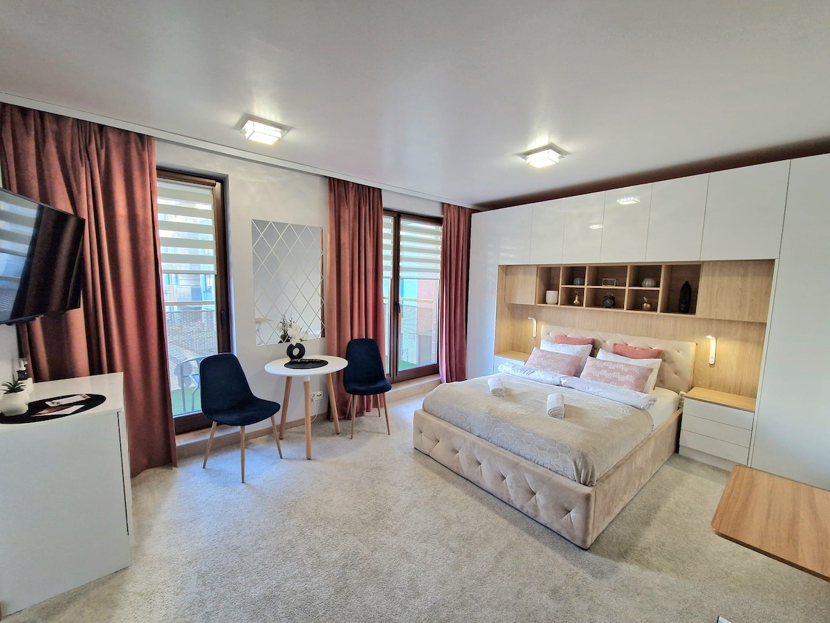A modern room features a spacious bed with soft bedding, complemented by decorative pillows. A small dining table with two chairs occupies one corner. Natural light enters through large windows framed by elegant curtains, providing a bright and open atmosphere.