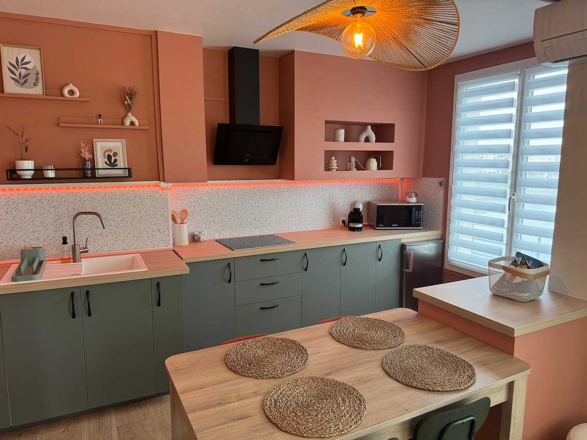 A modern kitchen area is illustrated with earthy tones, featuring green cabinetry and a light wood countertop. An artful light fixture hangs above a dining table set with woven placemats. Decorative shelving is visible, displaying plants and tableware, enhancing the inviting ambiance of the space.