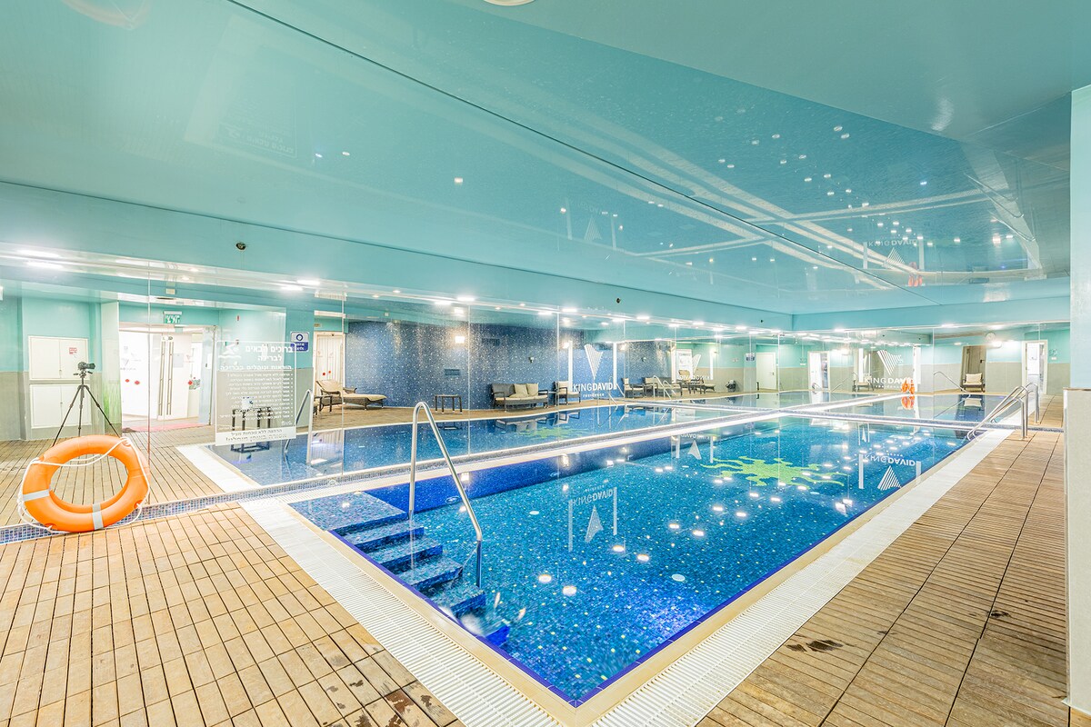 An indoor swimming pool features turquoise water reflecting the ceiling lights. Surrounding the pool, lounge chairs are positioned for relaxation, and a safety buoy is visible nearby. The area is decorated with blue tiles that add to the tranquil ambiance.