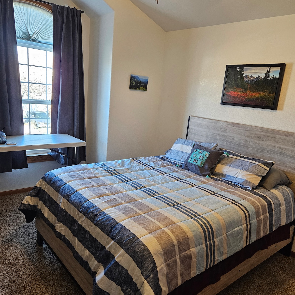 The spacious bedroom features a queen bed with a quilted striped coverlet. A window adorned with curtains allows natural light to fill the room. A small desk and chair are positioned beside the window, providing a workspace. A mountain-themed artwork hangs on the wall.
