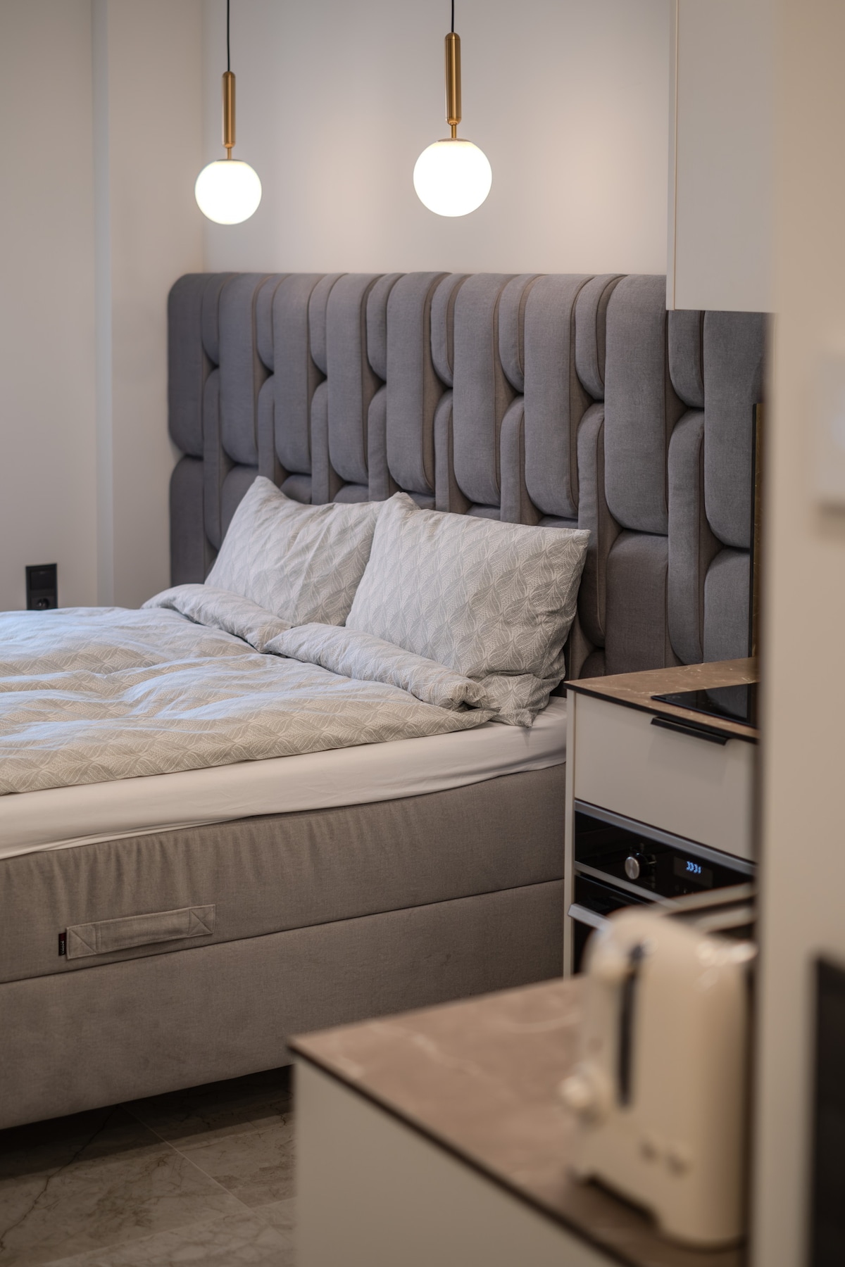 A modern bedroom features a plush grey upholstered headboard above a neatly made spacious bed with decorative pillows. Soft lighting is provided by two pendant lights overhead. A compact kitchenette is visible in the background with a kettle and toaster, enhancing the functionality of the space.