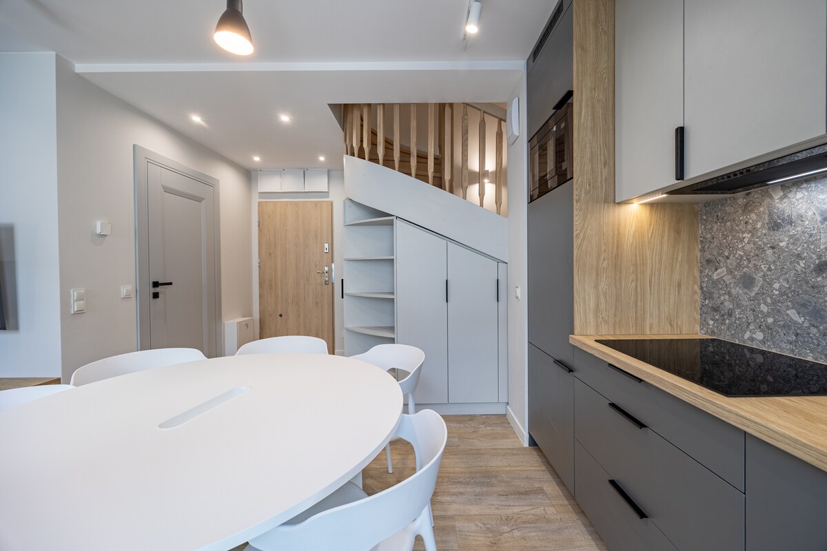 A dining area features a round white table surrounded by six chairs. A staircase with wooden steps leads to the upper level. The kitchen showcases modern cabinetry with a contrasting dark countertop, creating a sleek and functional space.