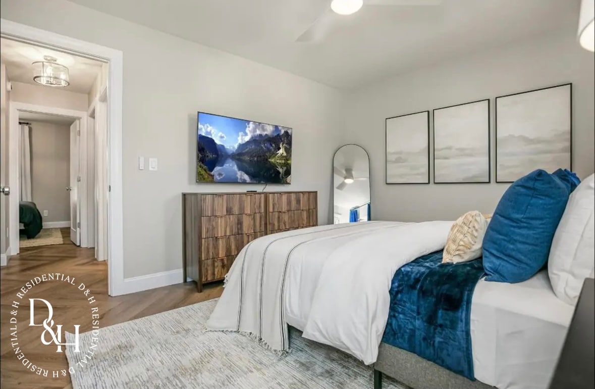 A well-appointed sleeping area is featured, showcasing a spacious bed made with crisp white linens and a soft throw blanket in blue. A wooden dresser with subtle texture stands against the wall, accompanied by a flat-screen television mounted above. Three framed artworks add interest to the neutral-toned walls.