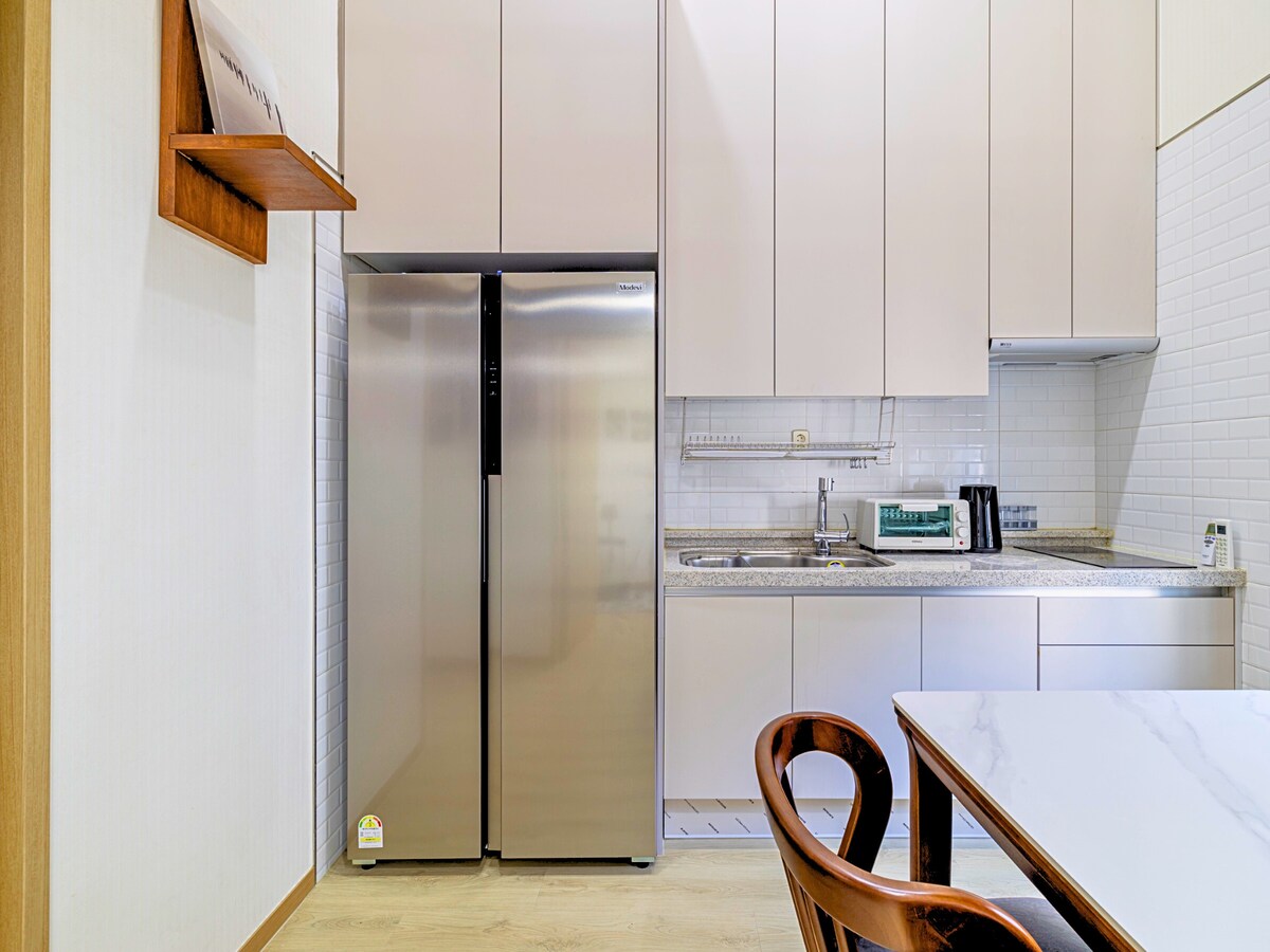 A modern kitchen features sleek cabinetry and a spacious countertop. The stainless steel refrigerator stands beside a stainless steel sink, with essential appliances like a microwave and electric kettle placed on the counter. A dining table with wooden chairs is positioned in the foreground.