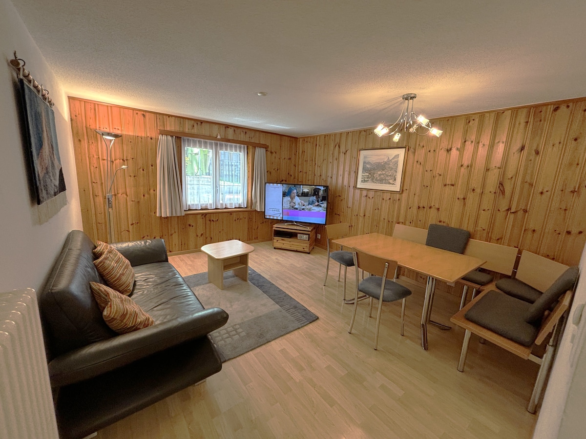 A living area features wooden paneling and soft lighting. A large flat-screen television sits on a cabinet, while a comfortable sofa and a coffee table provide seating. A dining table with chairs is positioned nearby, with natural light filtering through the windows.