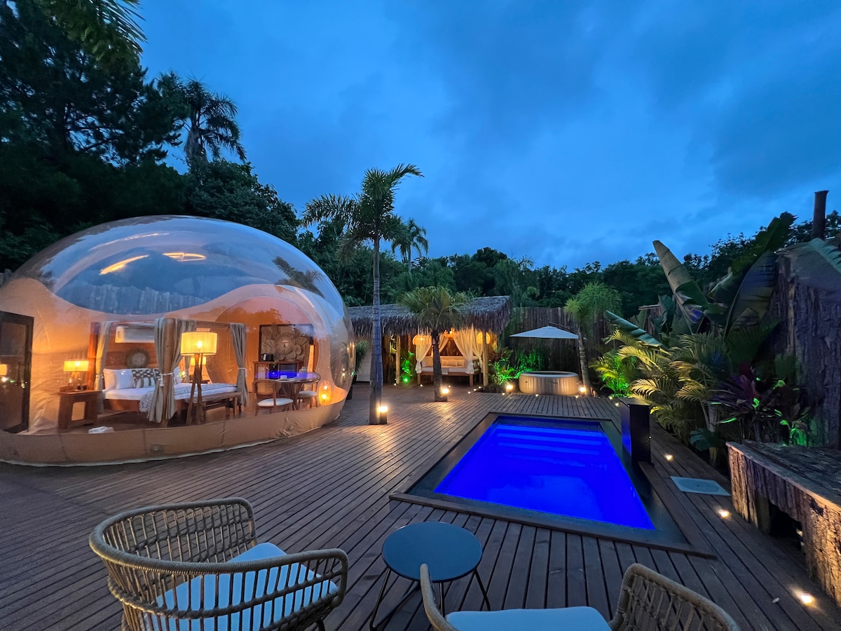 A bubble accommodation is featured on a wooden deck, surrounded by lush tropical vegetation. A private pool in a vivid blue reflects the evening sky, while soft lighting enhances the inviting ambiance. Comfortable seating is provided by decorative chairs and a small table.