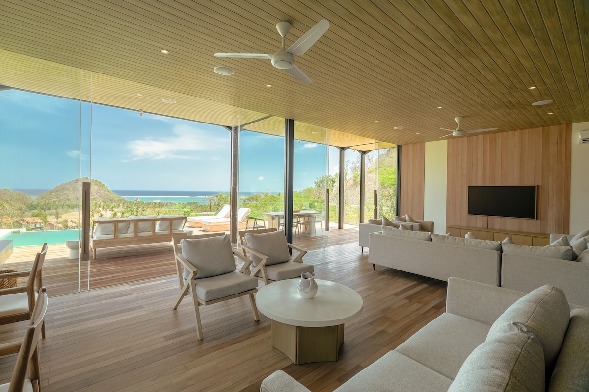 The spacious lounge area features large floor-to-ceiling glass walls that provide a panoramic view of the surrounding forest and sea. Comfortable seating arrangements include a light-colored sofa and several armchairs, with a central coffee table and a wall-mounted TV visible.