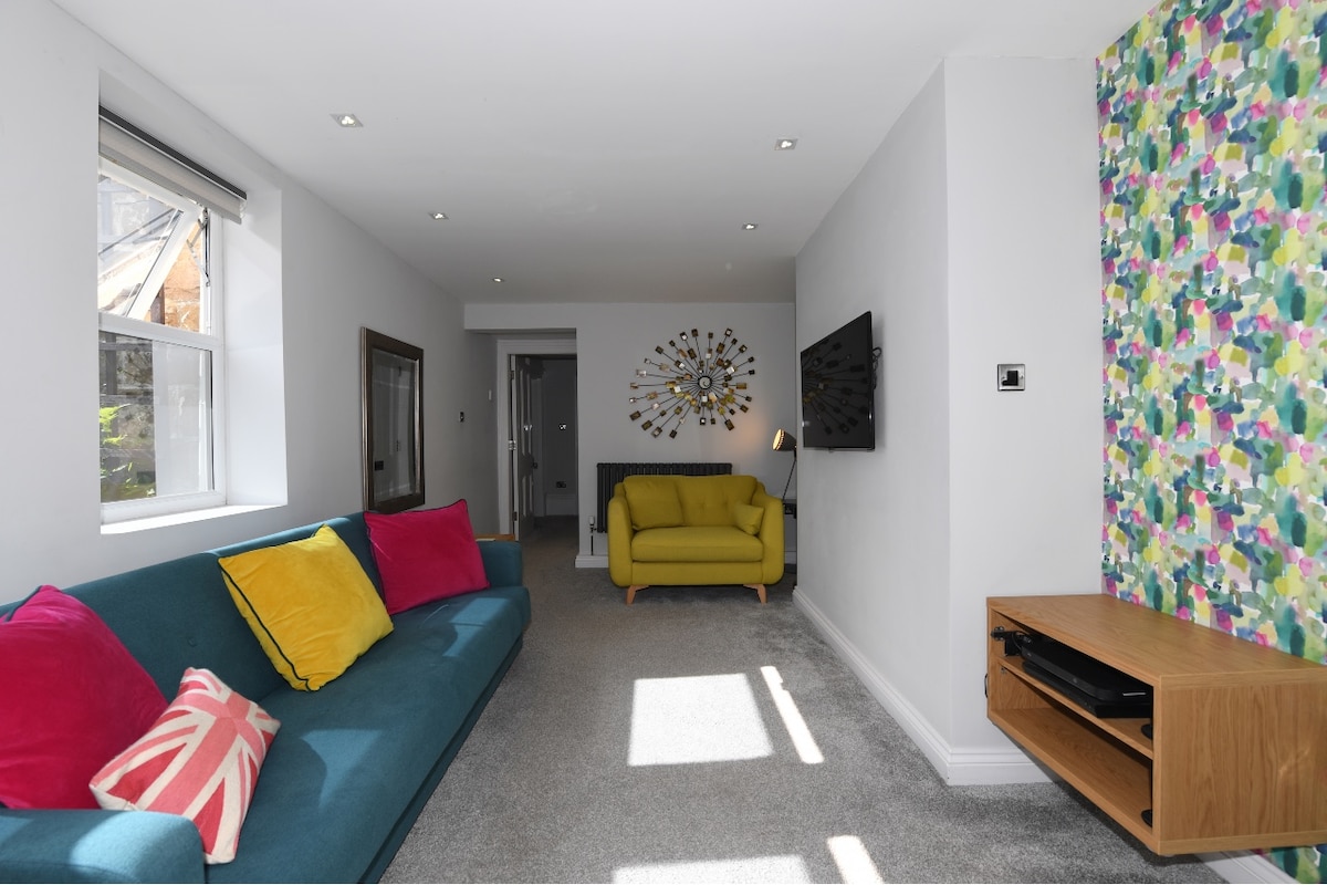 The spacious lounge area features a vibrant blue sofa adorned with colorful cushions, complemented by a mustard yellow armchair. A wall-mounted TV is positioned above a wooden media console. Natural light filters through the windows, creating a warm ambiance.