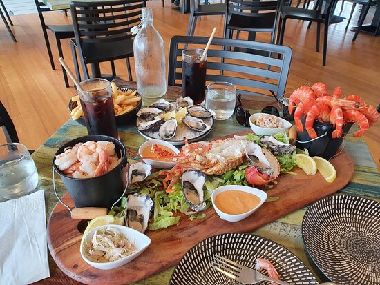 A wooden serving platter showcases a variety of seafood, including shrimp, oysters, and crab. Accompanied by dipping sauces and garnished with lettuce and lemon wedges, the arrangement is surrounded by drinks in glassware on a table with modern black chairs in a well-lit dining area.
