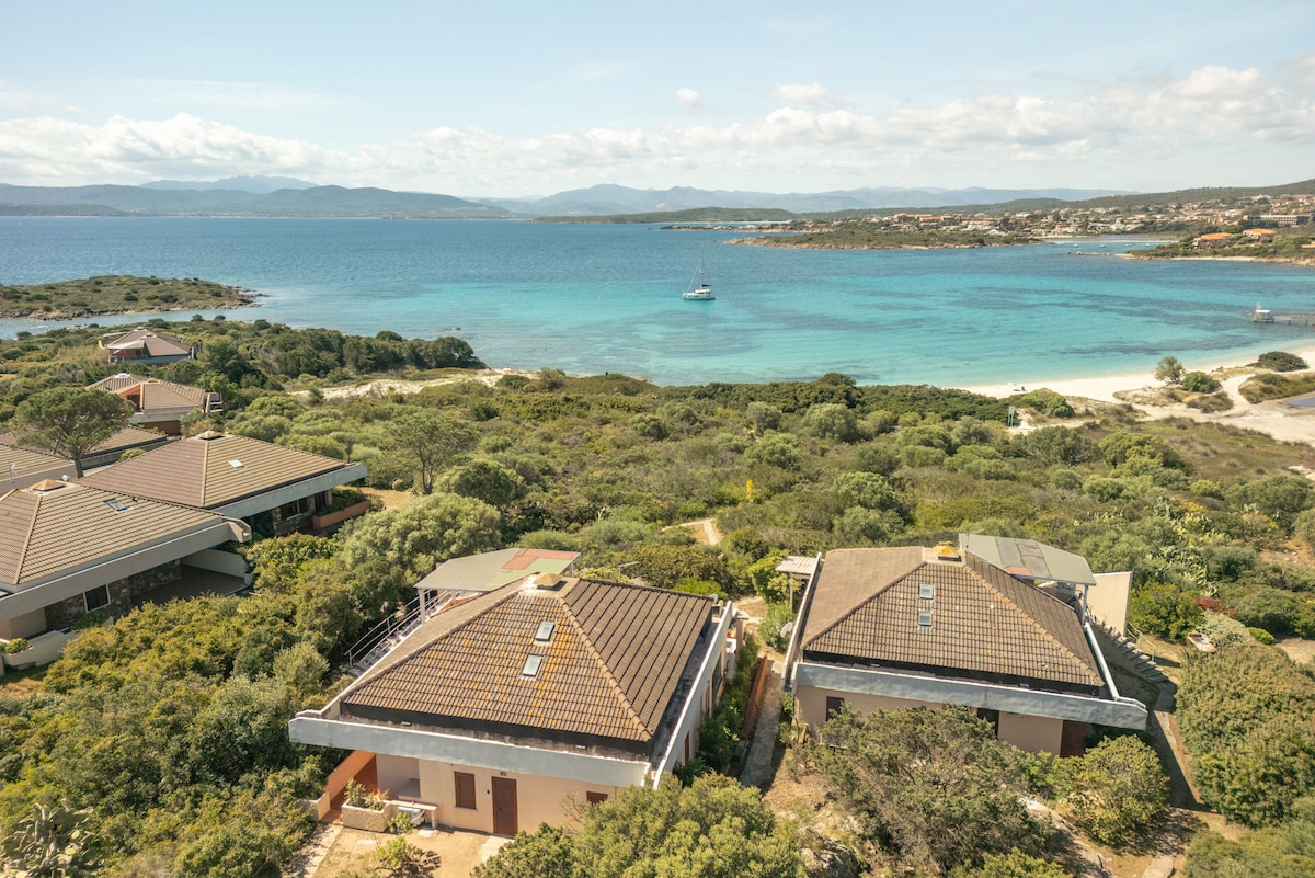 The image captures a scenic coastal view, showcasing a serene blue sea extending towards distant hills. Several structures with terracotta roofs are nestled within verdant vegetation, contributing to the natural, tranquil environment. A small boat can be seen in the water, adding a sense of exploration.