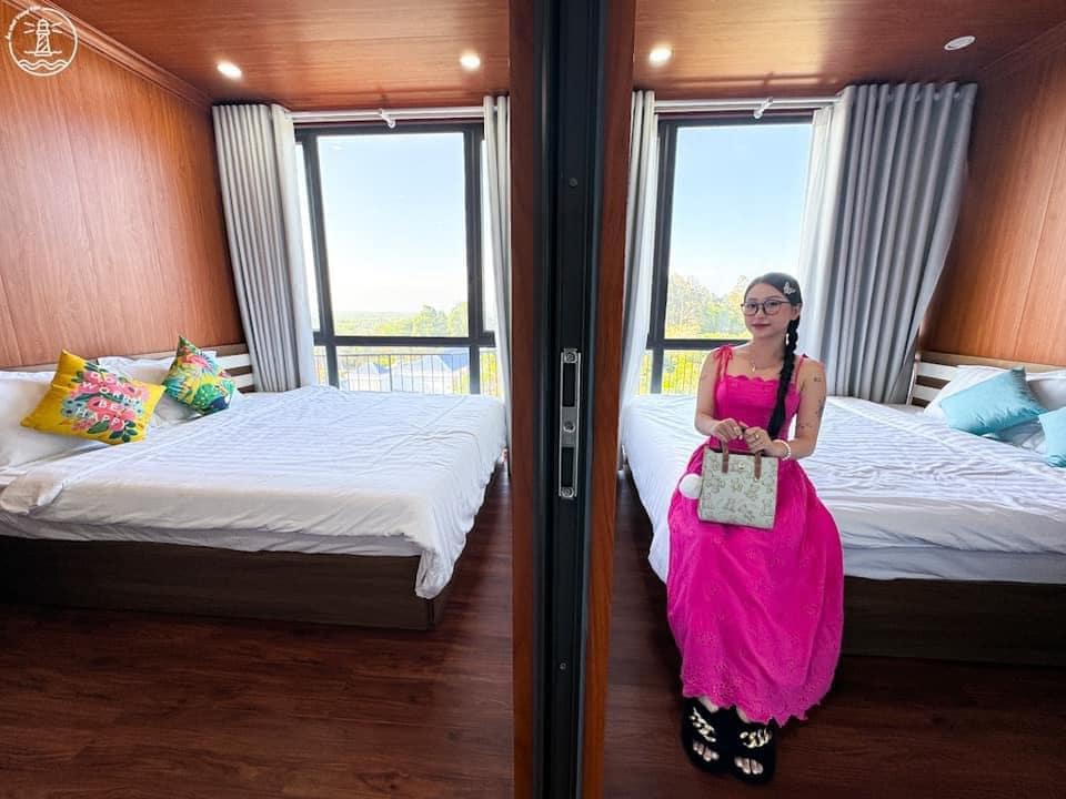 A bright and spacious room is presented, featuring two large beds adorned with white bedding. Large windows provide ample natural light, while vibrant decorative pillows add a pop of color. Neutral-toned wooden walls create a warm ambiance, enhancing the inviting environment.