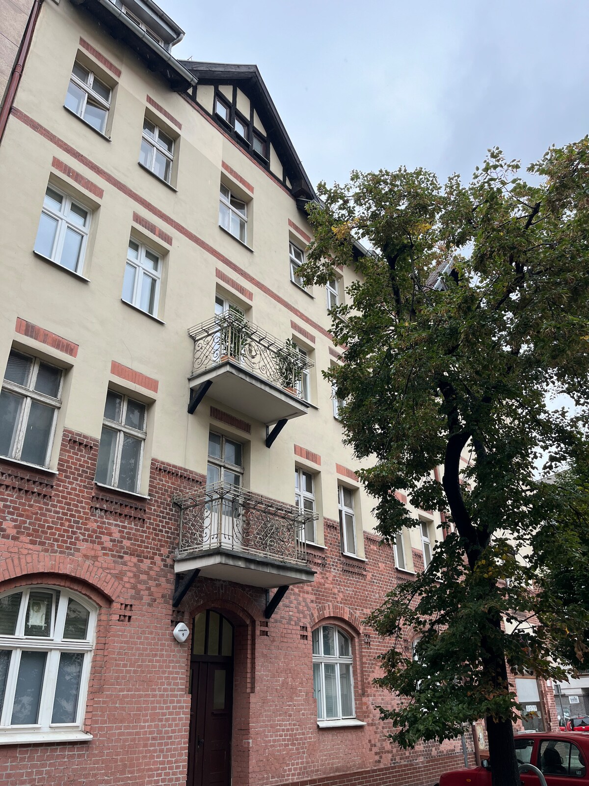 A historic building features a combination of brick and stucco facades, highlighted by balconies adorned with wrought iron railings. Tall trees flank the entrance, while multiple large windows allow for natural light to fill the interior spaces.