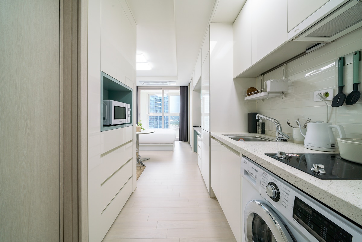 A bright kitchen is featured, with sleek white cabinetry and modern appliances, including a microwave and a washing machine. A window allows natural light to fill the space, highlighting the neutral tones of the flooring and countertops.