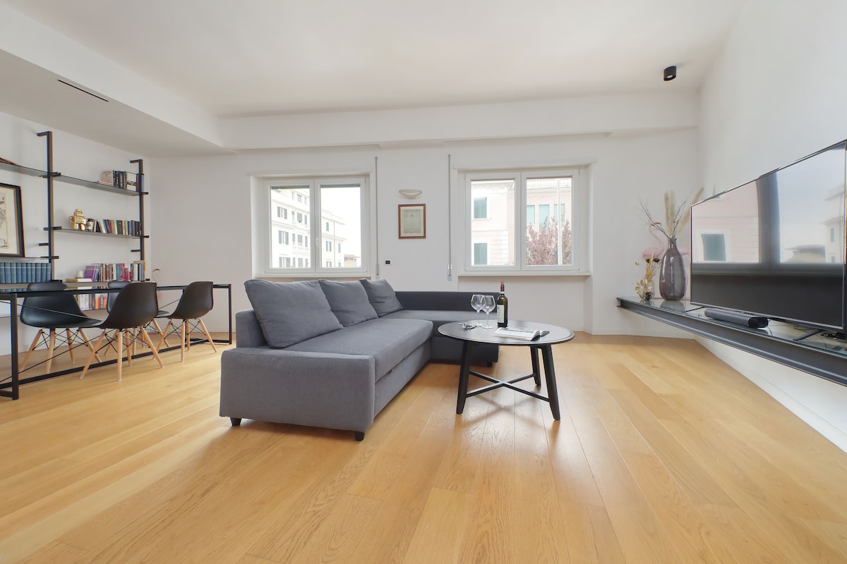 A spacious living area is presented, featuring a comfortable gray sofa and a modern coffee table. Natural light enters through two large windows, highlighting the warm wood flooring. A stylish bookshelf and dining area with chairs are visible in the background, enhancing the sense of openness.