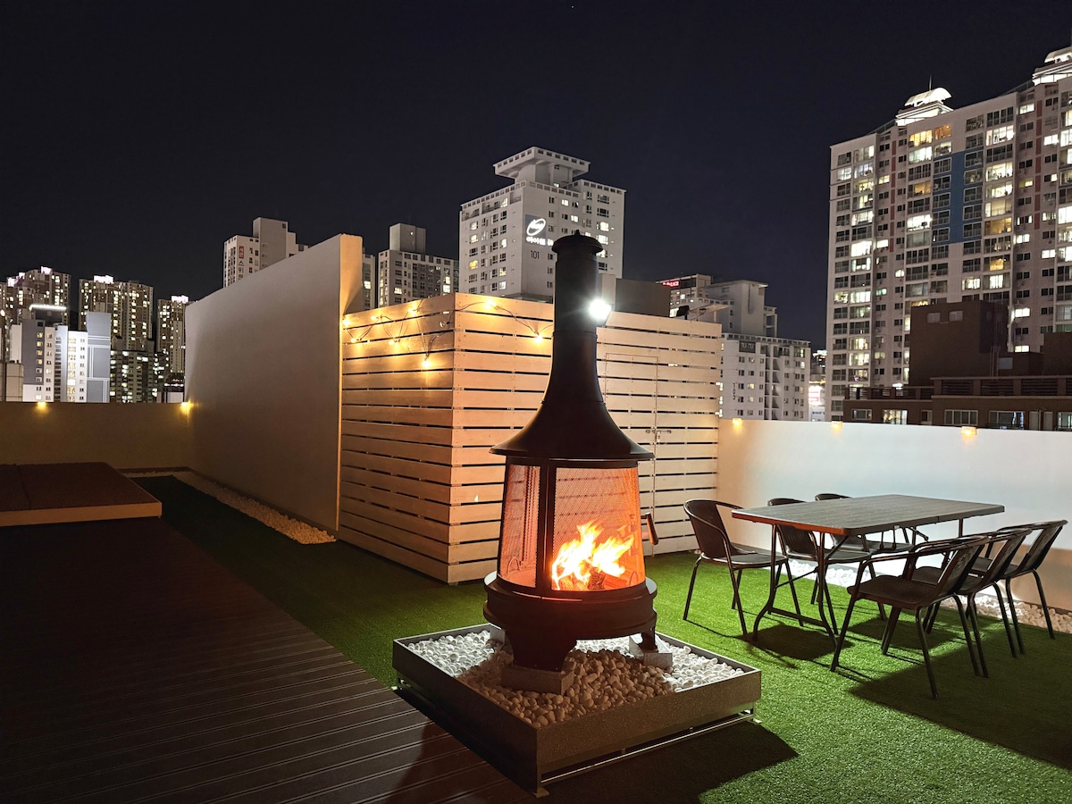 An open rooftop garden features a central fireplace surrounded by gravel. Four chairs and a table are positioned nearby, with bright city lights visible in the background. The area is designed with green synthetic grass and illuminated by soft outdoor lighting.