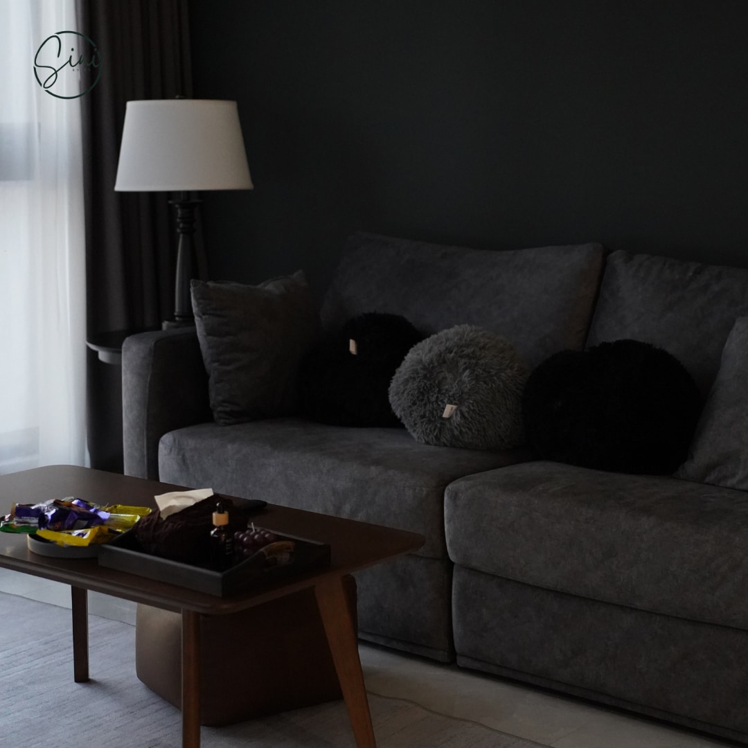 A modern living area features a spacious gray sofa adorned with textured black cushions. A low wooden coffee table is placed in front, displaying an assortment of snacks. A floor lamp stands to one side, providing ambient lighting.