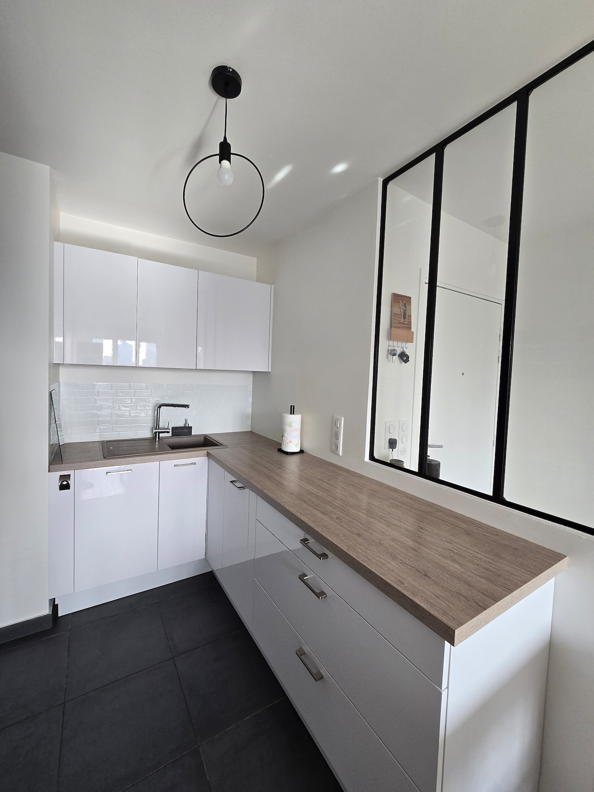 A modern kitchen space features sleek white cabinetry and a wooden countertop. A sink is integrated into the design, and a large mirror adds depth to the room. The floor is tiled in dark grey, complementing the overall aesthetic.