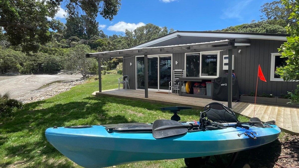 A kayak rests on the grass in front of a modern exterior of a dwelling with large windows. A wooden deck extends outwards, providing access to the beach. Nearby, a shaded area offers seating, contributing to a relaxed atmosphere surrounded by greenery.