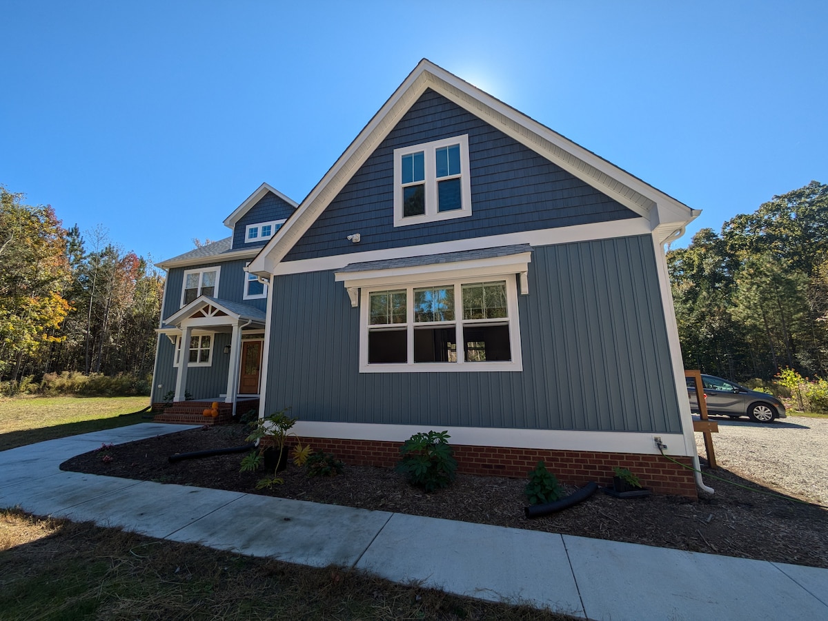 The exterior of a modern two-story house is shown, featuring a combination of blue siding and white trim. The property is surrounded by a landscaped area with small plants. A driveway is visible leading to a parked vehicle, framed by trees in the background.