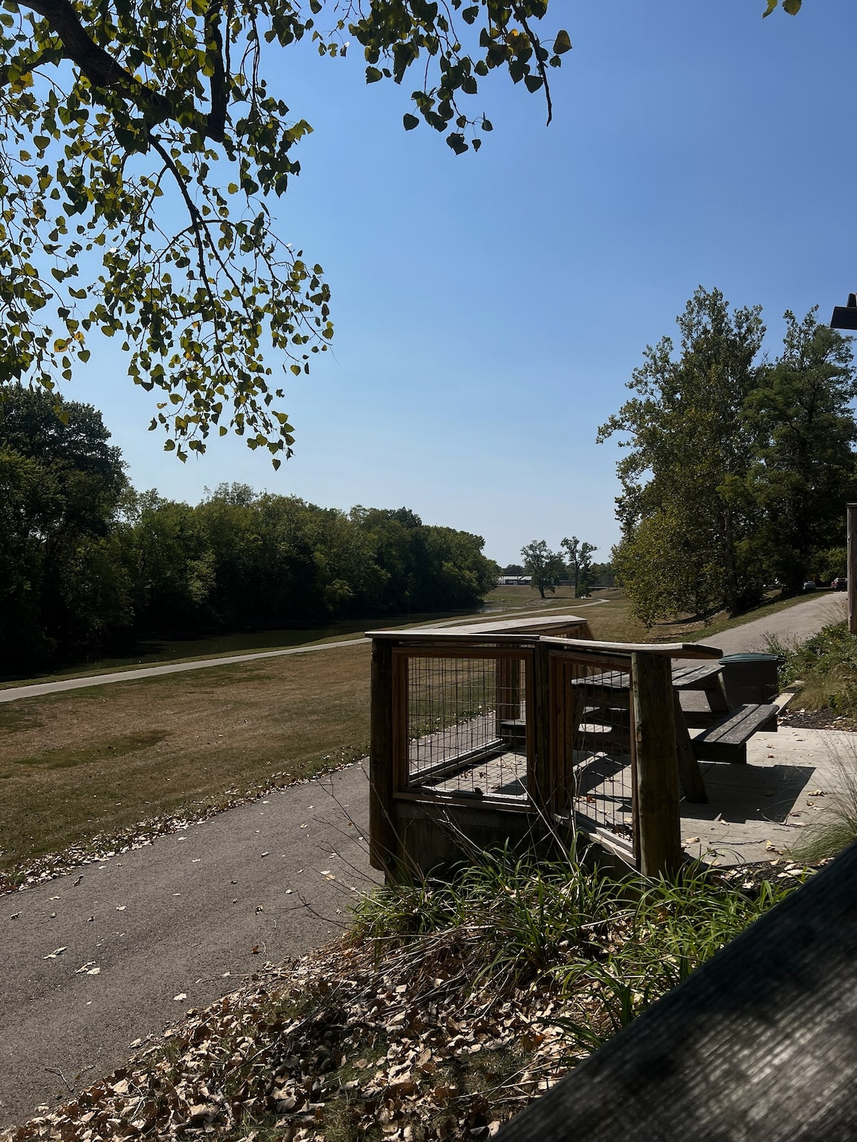 A wooden deck with a railing is shown, offering seating for outdoor dining. Nearby, a pathway leads into a green, open space bordered by trees, creating a serene environment under a clear blue sky.