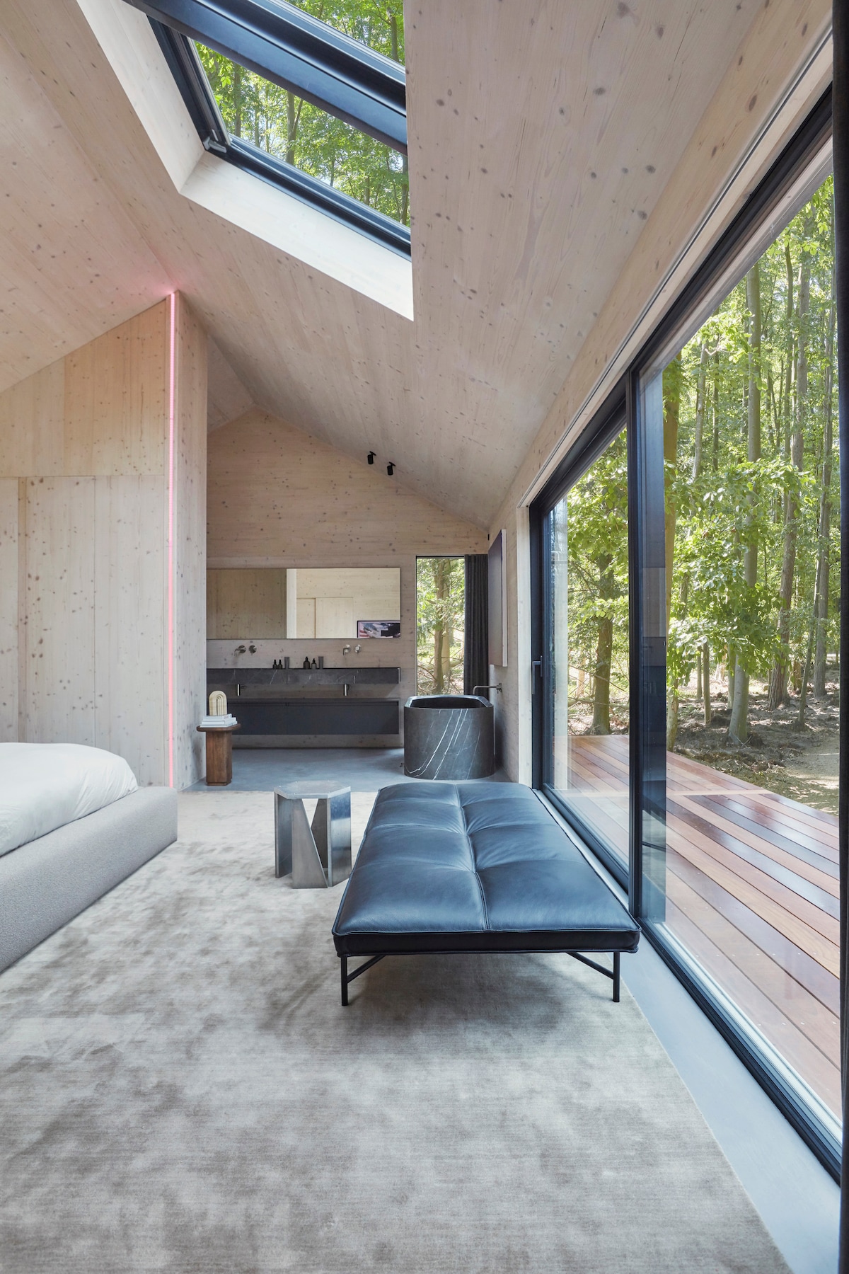 A cozy living area features a dark upholstered bench positioned on a plush rug. Large windows invite natural light, offering views of the surrounding forest. The room’s wooden walls and ceiling contribute to a warm aesthetic, while a modern television setup is visible in the background.