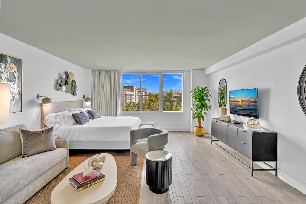 A bright, spacious studio suite features a king-sized bed with crisp linens and a cozy seating area. Large windows provide natural light and views of the surrounding landscaping. A sleek TV is mounted on the wall opposite the bed, complementing the contemporary décor.