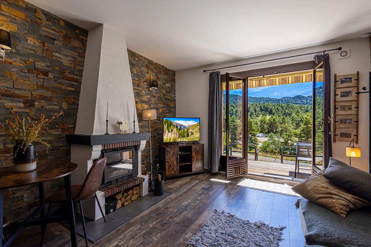 A cozy living area features a stone fireplace and a wooden table with a chair. Natural light floods in through the open balcony doors, which provide a view of the surrounding mountains. A television and decorative elements enhance the inviting atmosphere.