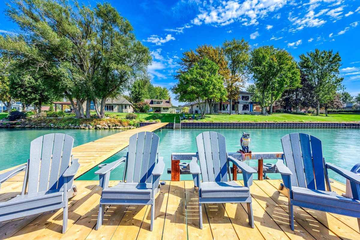 A wooden dock extends over the water, featuring several grey Adirondack chairs facing the serene river. Lush trees and a grassy area create a natural backdrop, while a small building is visible on the opposite bank, providing a charming view.