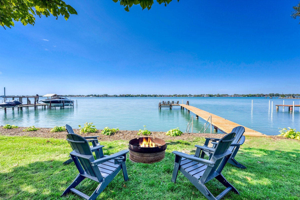 Two Adirondack chairs are positioned around a fire pit on a grassy area overlooking the river. A wooden dock extends into the water, with boats visible in the distance. The clear blue sky reflects peacefulness in this serene outdoor setting.