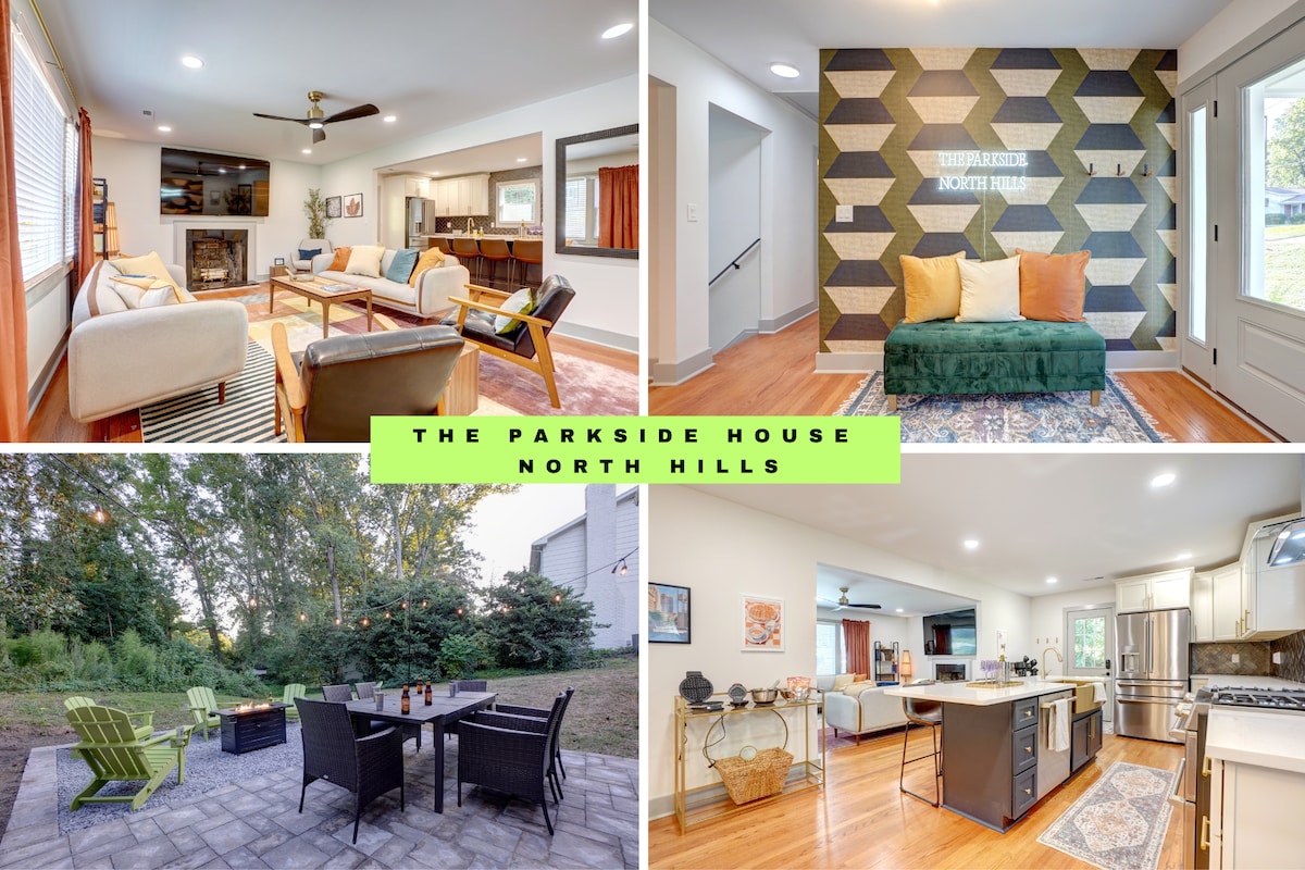 A bright living room features ample seating arranged around a large TV, illuminated by natural light from windows. The kitchen is visible in the background, showcasing modern appliances. Outside, a patio area with lounge chairs and a fire pit is surrounded by natural greenery.