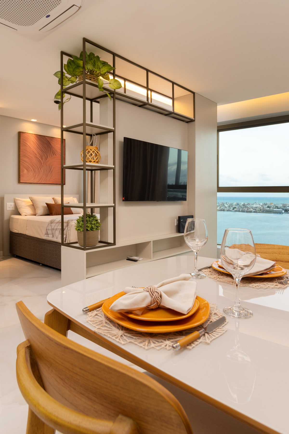 A stylish dining area is visible, featuring a modern table set with elegant tableware. Behind it, a wall shelf adorned with greenery adds a touch of vibrancy. A large window offers a panoramic view of the surroundings, while a cozy sleeping space is framed in the background.