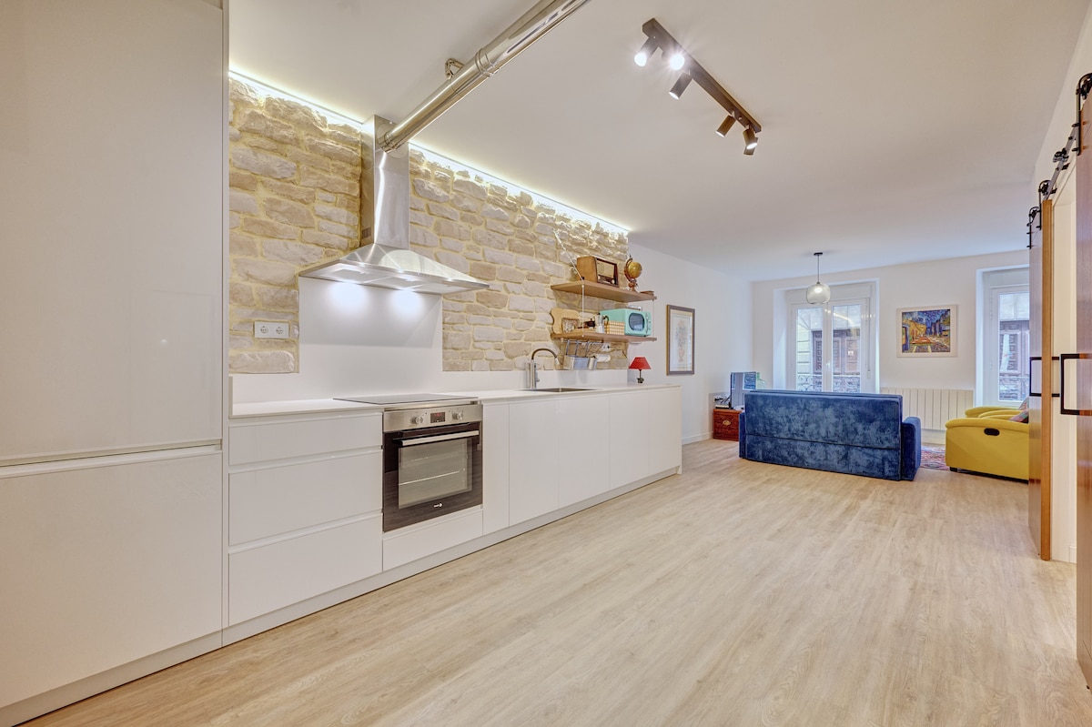 A modern kitchen features sleek cabinetry and an integrated oven. A textured stone wall adds character to the space. The open layout allows for easy movement, leading to a cozy living area with a blue sofa and a vibrant yellow chair, creating a welcoming environment.