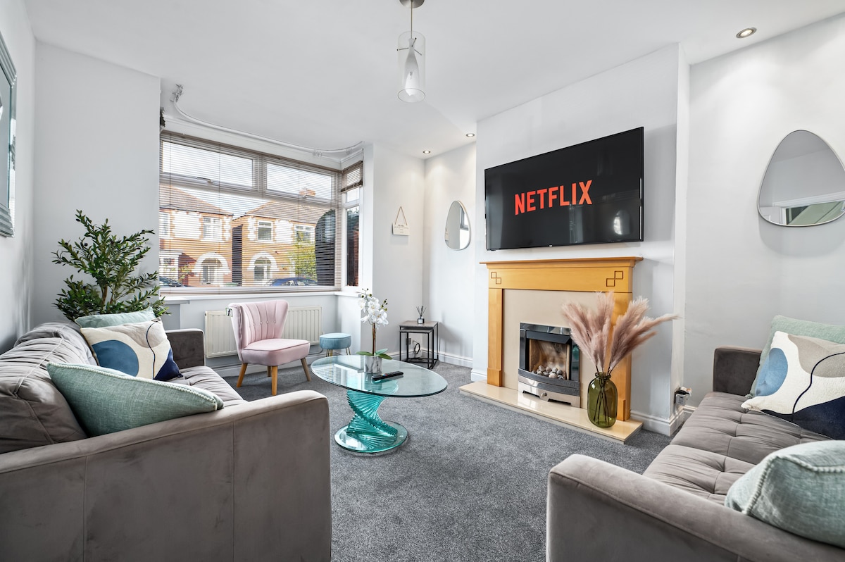 A modern living area features two grey sofas adorned with colorful cushions, situated on plush grey carpeting. A glass coffee table rests in the center, while a fireplace adds warmth. A large television mounted on the wall displays the Netflix logo, and natural light enters through a large window.