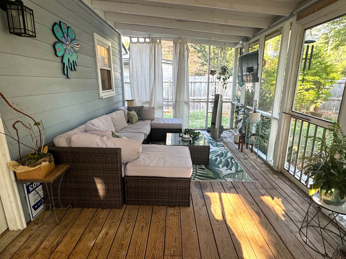 A screened-in porch is featured with comfortable wicker seating arranged around a coffee table. Natural light floods the space, highlighting the wooden floors and green foliage decor. Sheer curtains gently billow, providing a serene view of the backyard through the screens.