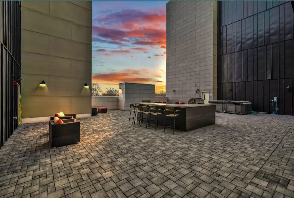 A rooftop terrace features paved flooring and a seating area with a couch and bar stools around a large rectangular table. A grill and hot tub are also evident. The setting sun casts a warm glow over the space, contrasting with the building exteriors.