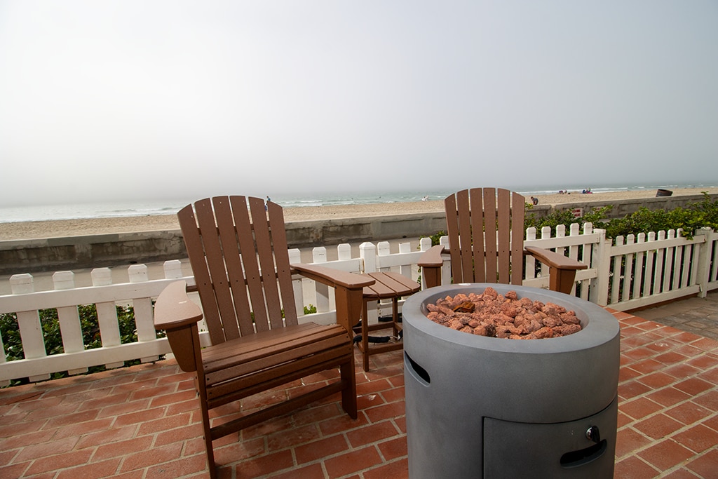Two Adirondack chairs are positioned near a circular propane firepit on a spacious brick patio. The serene beach view extends beyond a white picket fence, with gentle waves lapping at the shore. A misty atmosphere adds a calming ambiance to the outdoor space.