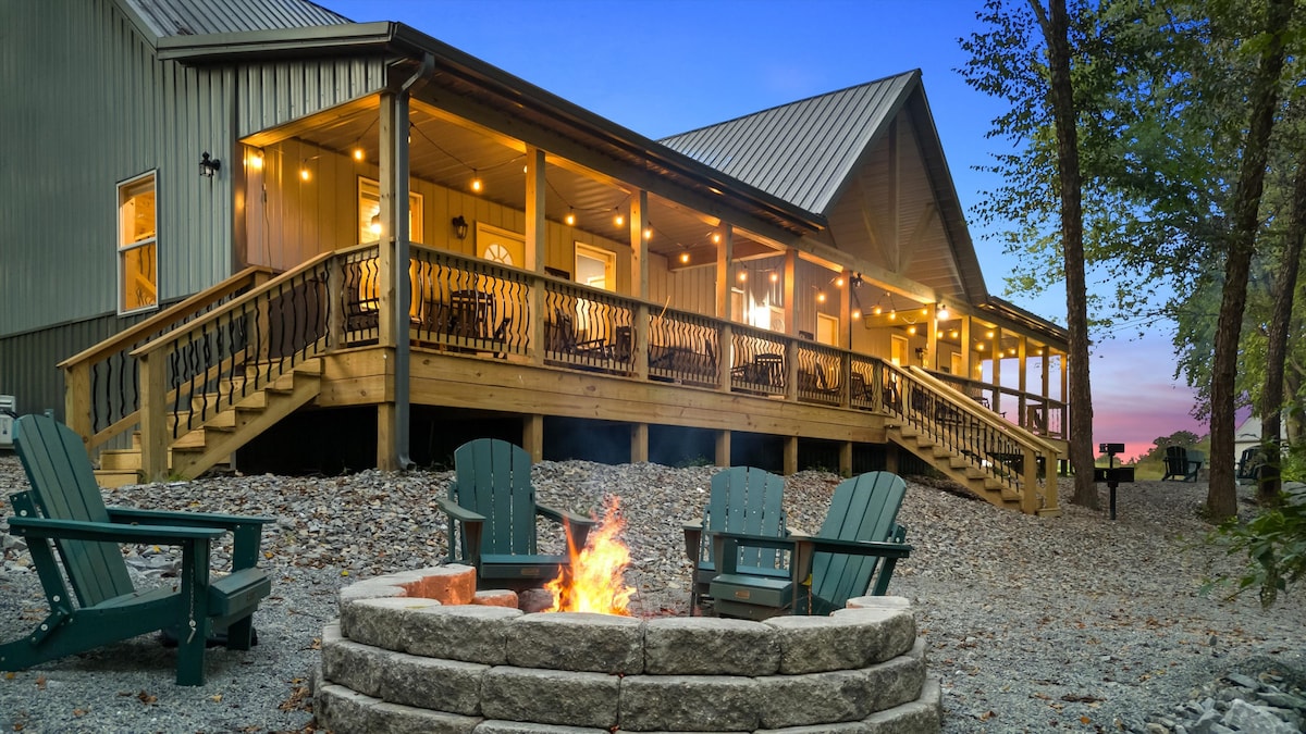 A spacious cabin porch features multiple rocking chairs, inviting relaxation while enjoying riverfront views. A circular fire pit surrounded by green Adirondack chairs is located on the stone pathway, providing a cozy gathering space. Warm lighting enhances the cabin's exterior at dusk.