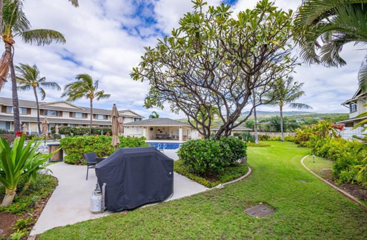 A landscaped outdoor area features a lush green lawn, surrounded by tropical plants and palm trees. A barbecue grill is placed on a concrete patio, with a large tree providing shade. In the background, a community pool reflects the sky, enhancing the tranquil atmosphere.