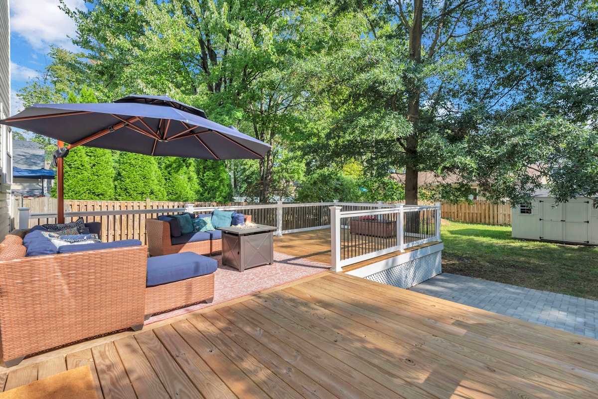 An outdoor deck area is shown featuring a shaded seating arrangement under a large umbrella. Comfortable seating is provided with plush cushions, and a stylish coffee table occupies the center. Lush greenery surrounds the space, enhancing the peaceful atmosphere.