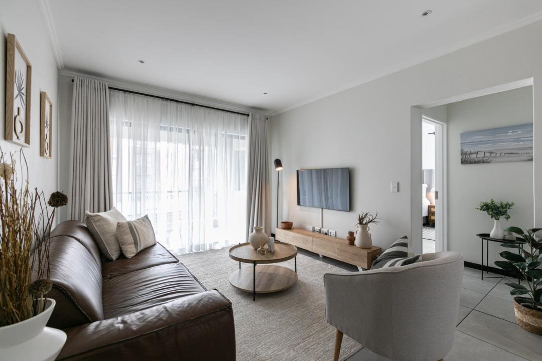 The living area features a comfortable leather couch and a stylish occasional chair, positioned around a round coffee table. A smart TV is mounted on the wall, while sheer curtains frame large windows, allowing natural light to fill the space. A neutral-toned rug complements the minimalist decor.