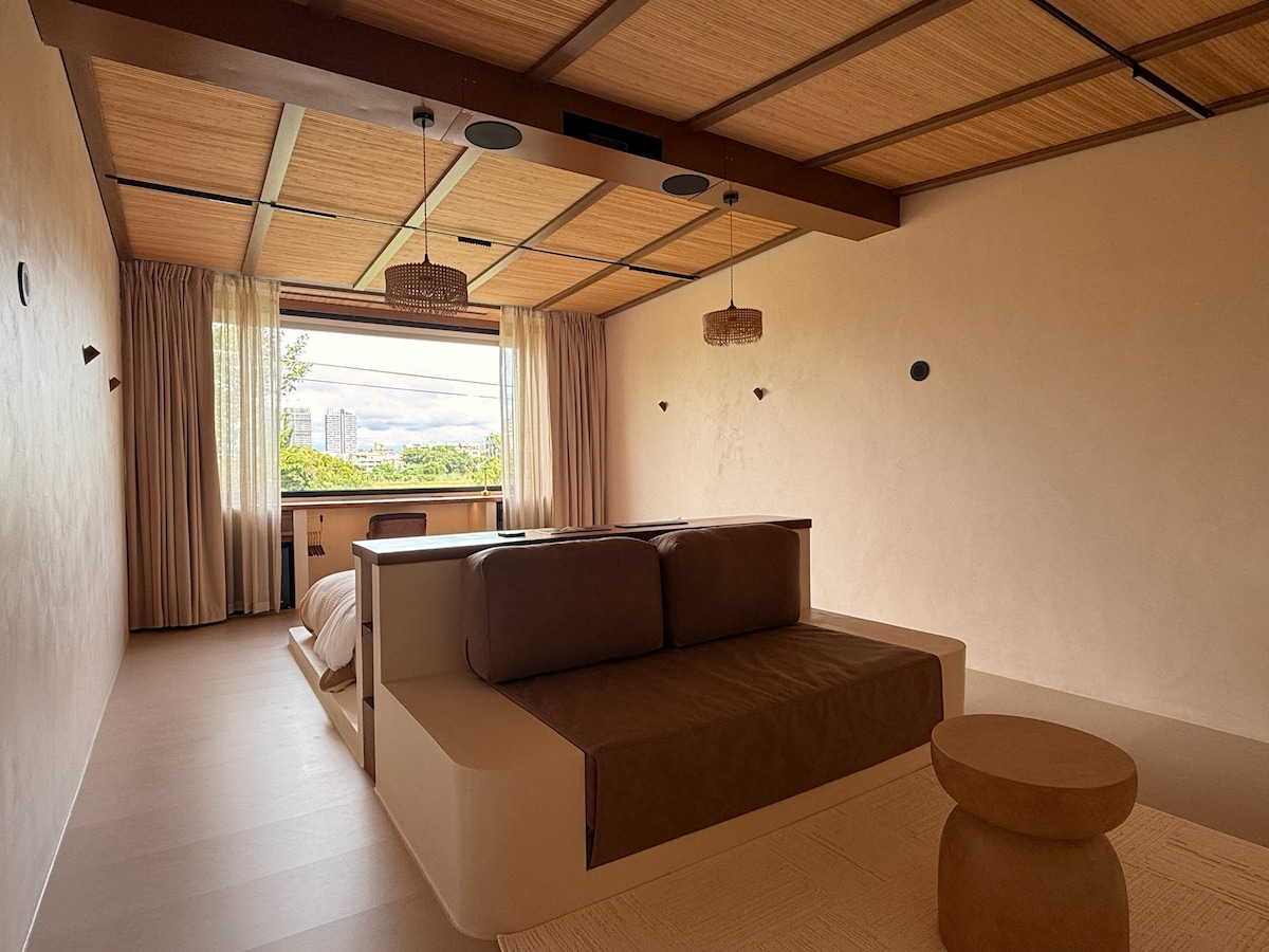 A spacious room features a large window with views of the surrounding greenery. The warm wooden ceiling contrasts with the light-colored walls. A minimalist sitting area includes a sofa and a round coffee table, creating a serene and inviting space for relaxation.