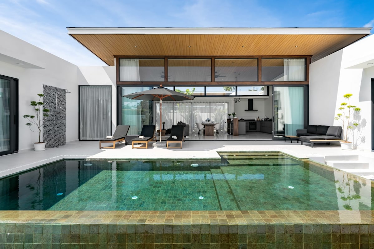 A modern villa features an outdoor area with a private pool, sun loungers, and an umbrella. The interior design includes large glass windows, allowing natural light to fill the space. A dining area is visible in the background, offering a blend of indoor and outdoor living.